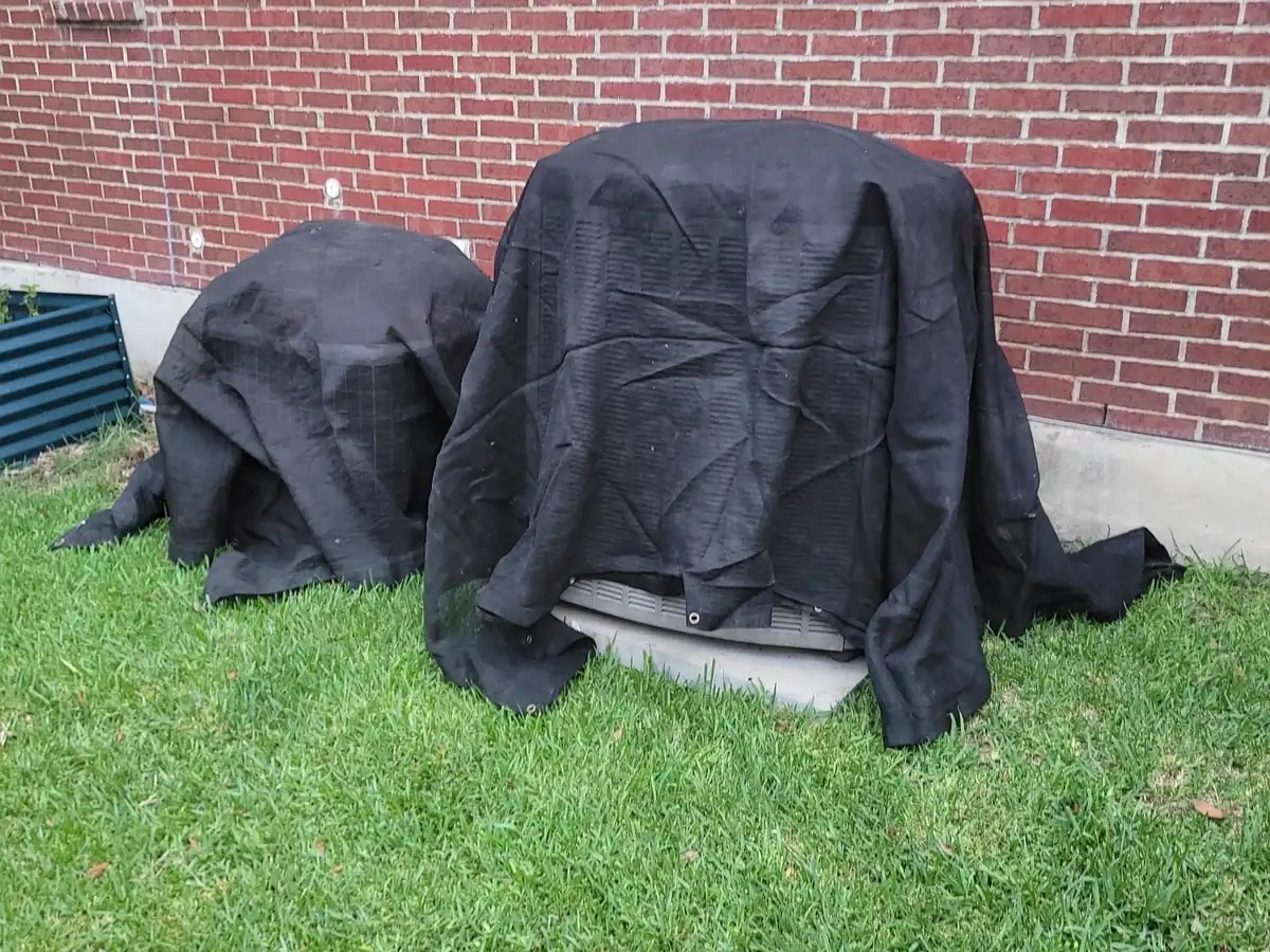 Two air conditioning units beside a brick house covered in black mesh to protect them during a roof replacement